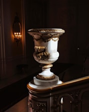 A large, ornate marble urn is placed on a decorative staircase in a dimly lit interior. The urn is adorned with intricate gold embellishments and floral motifs. To the left, a wall-mounted candle holder with multiple lit candles provides a warm glow, illuminating the ornate details of the architecture.