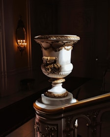 A large, ornate marble urn is placed on a decorative staircase in a dimly lit interior. The urn is adorned with intricate gold embellishments and floral motifs. To the left, a wall-mounted candle holder with multiple lit candles provides a warm glow, illuminating the ornate details of the architecture.