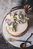 A round cake adorned with a cascade of white and purple flowers is placed on a marble surface. The cake sits on a golden tray, and nearby are elegant gold-tone cutlery and a match holder filled with decorative matches.
