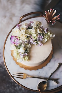 A round cake adorned with a cascade of white and purple flowers is placed on a marble surface. The cake sits on a golden tray, and nearby are elegant gold-tone cutlery and a match holder filled with decorative matches.