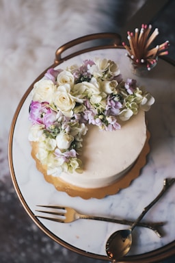 A round cake adorned with a cascade of white and purple flowers is placed on a marble surface. The cake sits on a golden tray, and nearby are elegant gold-tone cutlery and a match holder filled with decorative matches.