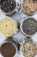Several glass jars filled with different types of loose leaf teas are arranged on a light surface. Each jar contains a distinct type of tea, with varying colors and textures, including chamomile, black tea, and herbal blends.