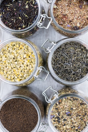 Several glass jars filled with different types of loose leaf teas are arranged on a light surface. Each jar contains a distinct type of tea, with varying colors and textures, including chamomile, black tea, and herbal blends.