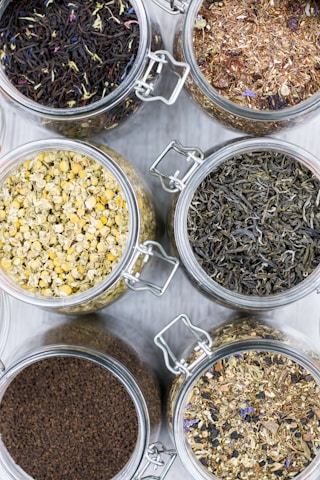 Several glass jars filled with different types of loose leaf teas are arranged on a light surface. Each jar contains a distinct type of tea, with varying colors and textures, including chamomile, black tea, and herbal blends.