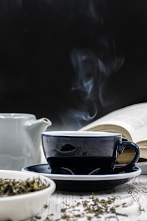 A steaming cup of tea sits on a saucer in front of an open book. A teapot is visible in the background, and loose tea leaves are scattered in a white dish on the table. The scene suggests a calm and quiet atmosphere.
