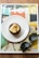 A cupcake with creamy frosting sits on a white plate, which is placed on a magazine titled 'frieze'. Sunglasses rest beside the plate, adding a casual and stylish touch to the composition. The magazine's cover features an abstract artwork with geometric patterns and vibrant colors.
