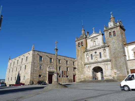 A historic stone cathedral with intricate architectural details stands prominently under a clear blue sky. The facade features tall towers, decorative statues, and an arched entrance. Adjacent to the cathedral is a large, rectangular building with many windows. In the foreground, a stone cross stands atop a pedestal. Several vehicles are parked nearby, including a white van.