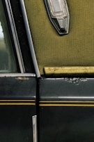 A close-up view of an old car door, featuring a worn textured green interior fabric, a metallic trim, and a vintage light fixture. The exterior is dark with yellow pinstripes and shows signs of age with rust and scratches.