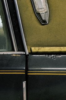 A close-up view of an old car door, featuring a worn textured green interior fabric, a metallic trim, and a vintage light fixture. The exterior is dark with yellow pinstripes and shows signs of age with rust and scratches.