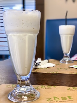 A close-up view of two tall, frothy milkshakes or smoothies served in clear glass cups. The glasses are placed on a wooden table with paper placemats labeled Zest Cafe & Restaurant. Napkins and cutlery are visible in the background.
