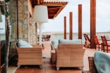An outdoor patio area with wicker furniture and light blue cushions. The setting includes a stone wall on the left and wooden columns supporting a roof structure. Multiple wooden chairs are arranged to face the view, and a large white floor lamp is positioned beside the furniture.