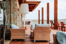 An outdoor patio area with wicker furniture and light blue cushions. The setting includes a stone wall on the left and wooden columns supporting a roof structure. Multiple wooden chairs are arranged to face the view, and a large white floor lamp is positioned beside the furniture.