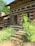 A rustic log cabin surrounded by lush greenery, featuring wooden shutters and a stone staircase leading to the entrance. The structure has a natural, earthy ambiance with rich wooden textures and a quaint charm.