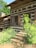 A rustic log cabin surrounded by lush greenery, featuring wooden shutters and a stone staircase leading to the entrance. The structure has a natural, earthy ambiance with rich wooden textures and a quaint charm.