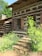 A rustic log cabin surrounded by lush greenery, featuring wooden shutters and a stone staircase leading to the entrance. The structure has a natural, earthy ambiance with rich wooden textures and a quaint charm.