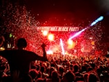 A vibrant outdoor party scene with a large crowd gathered in front of a stage. Bright lights and confetti fill the air, creating an energetic atmosphere. The stage is prominently labeled 'Capitol Hill Block Party' and colorful beams of light illuminate the night.
