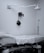 A clinical room with a medical examination bed covered in white sheets. A wall-mounted adjustable lamp is positioned above the bed. Medical equipment, including a blood pressure cuff, is mounted on the wall. The room is minimalistic and appears well-organized.
