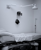 A clinical room with a medical examination bed covered in white sheets. A wall-mounted adjustable lamp is positioned above the bed. Medical equipment, including a blood pressure cuff, is mounted on the wall. The room is minimalistic and appears well-organized.