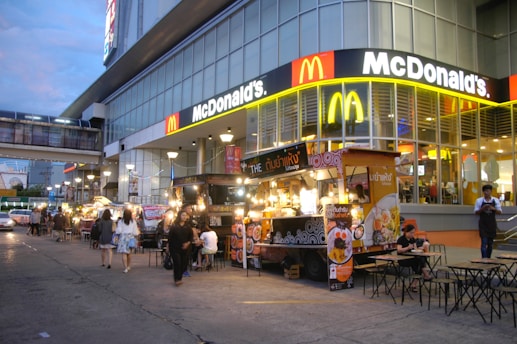 A busy street scene features a McDonald's with bright signage and a row of street food stalls offering various dishes. People are casually walking and dining, adding to the lively city atmosphere. The building's architecture is modern with large glass panels.