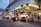 A busy street scene features a McDonald's with bright signage and a row of street food stalls offering various dishes. People are casually walking and dining, adding to the lively city atmosphere. The building's architecture is modern with large glass panels.