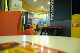 A fast-food restaurant interior with modern decor featuring a prominent red and yellow theme. There are several tables and chairs, with a group of people seated in the background having a conversation. Bright overhead lighting and large glass windows create an open atmosphere. The brand's logo and slogan are visible on the walls, and a sign advertising free Wi-Fi is displayed.