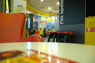 A fast-food restaurant interior with modern decor featuring a prominent red and yellow theme. There are several tables and chairs, with a group of people seated in the background having a conversation. Bright overhead lighting and large glass windows create an open atmosphere. The brand's logo and slogan are visible on the walls, and a sign advertising free Wi-Fi is displayed.