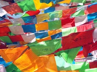 Colorful fabric flags with intricate designs and text are strung across each other in a vibrant display. The flags flutter gently in a breeze, catching light at different angles, creating a lively and bright atmosphere.