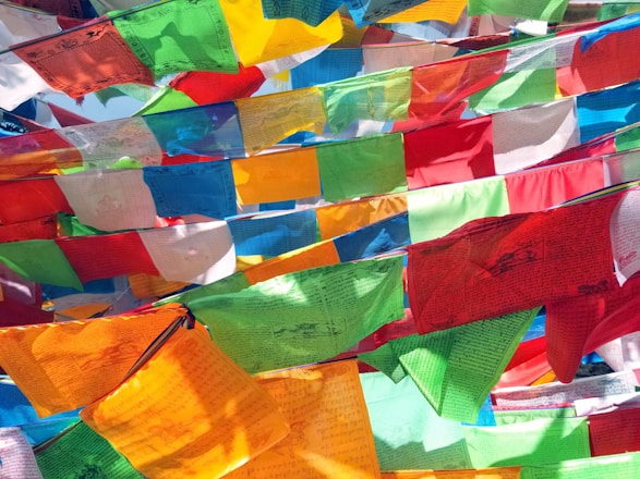 Colorful fabric flags with intricate designs and text are strung across each other in a vibrant display. The flags flutter gently in a breeze, catching light at different angles, creating a lively and bright atmosphere.