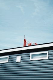 A Canadian flag waves atop a building with blue siding and small windows. The sky is clear and blue.