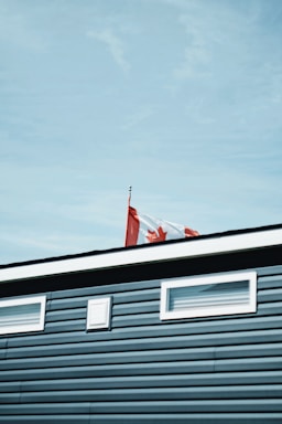 A Canadian flag waves atop a building with blue siding and small windows. The sky is clear and blue.