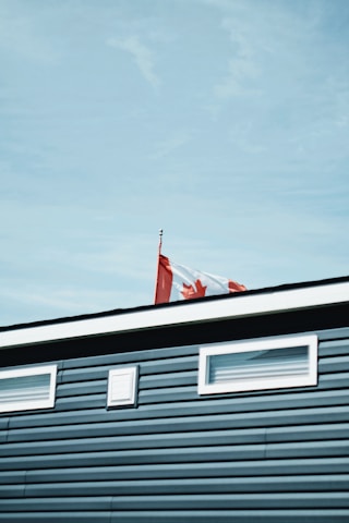 A Canadian flag waves atop a building with blue siding and small windows. The sky is clear and blue.