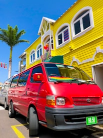 A vibrant scene featuring a bright red van parked in front of a two-story yellow wooden building. The building has white trim and arched windows, and there are colorful balloons visible at the entrance, suggesting a festive atmosphere. A tall palm tree is on the left side, with a clear blue sky above.
