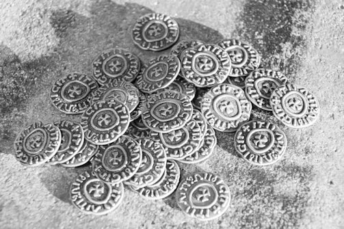 A pile of old-looking coins with intricate designs and inscriptions, scattered on a textured surface. Each coin features a star or cross-like symbol in the center, surrounded by circular text.