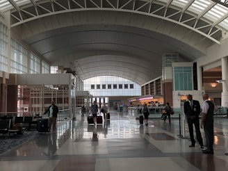 A spacious airport terminal with high arched ceilings and wide walkways, featuring several travelers rolling luggage. The area is brightly lit by natural light coming through large windows. Two people in uniforms are conversing on the side, while other passengers move toward check-in counters in the background. The sign above indicates 'Richmond'.