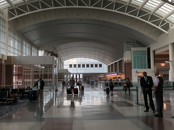 A spacious airport terminal with high arched ceilings and wide walkways, featuring several travelers rolling luggage. The area is brightly lit by natural light coming through large windows. Two people in uniforms are conversing on the side, while other passengers move toward check-in counters in the background. The sign above indicates 'Richmond'.