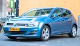 A blue Volkswagen car is parked on a paved street. The car has a license plate from the Netherlands, and the background includes a wooden barrier and some urban elements like buildings and signs.