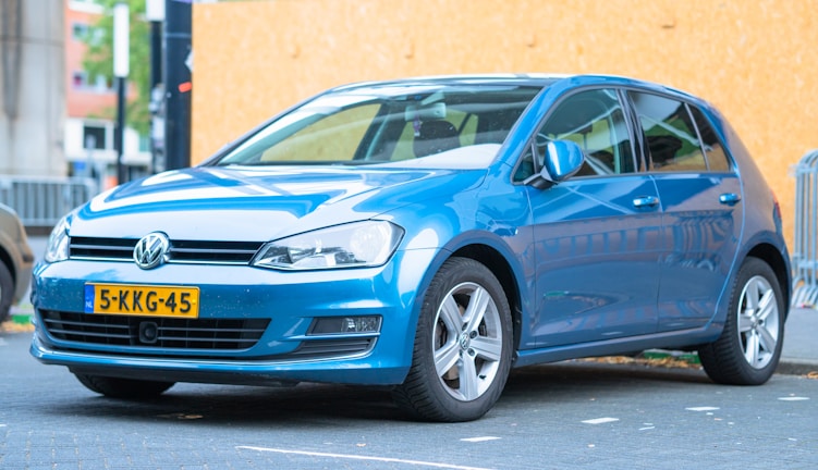 A blue Volkswagen car is parked on a paved street. The car has a license plate from the Netherlands, and the background includes a wooden barrier and some urban elements like buildings and signs.