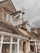 A quaint stone building with a vintage feel, featuring a prominent sign for 'The Goldsmithy Jewellers'. The facade includes rustic stone walls, traditional wooden window frames, and a sign stating 'Jewellery Designed & Made in Our Workshop'. The sky appears overcast, contributing to a cozy atmosphere.