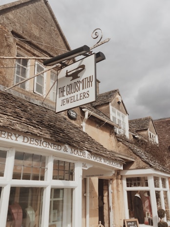 A quaint stone building with a vintage feel, featuring a prominent sign for 'The Goldsmithy Jewellers'. The facade includes rustic stone walls, traditional wooden window frames, and a sign stating 'Jewellery Designed & Made in Our Workshop'. The sky appears overcast, contributing to a cozy atmosphere.