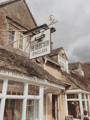 A quaint stone building with a vintage feel, featuring a prominent sign for 'The Goldsmithy Jewellers'. The facade includes rustic stone walls, traditional wooden window frames, and a sign stating 'Jewellery Designed & Made in Our Workshop'. The sky appears overcast, contributing to a cozy atmosphere.