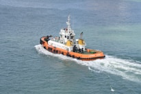 A tugboat with an orange hull navigates through calm waters, leaving a white wake in its path. The deck is equipped with various machinery and equipment, and several people are visible on board. The sea is a blend of deep blue and green hues, creating a serene and expansive backdrop.