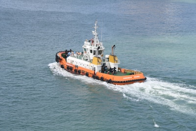 A tugboat with an orange hull navigates through calm waters, leaving a white wake in its path. The deck is equipped with various machinery and equipment, and several people are visible on board. The sea is a blend of deep blue and green hues, creating a serene and expansive backdrop.