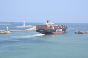 A large container ship labeled NYK LINE is navigating through a channel with breakwaters on either side. The ship is carrying numerous colorful shipping containers stacked on its deck. In the background, there are two lighthouses and another vessel can be seen on the open sea. The sky is clear and the water is calm, suggesting a peaceful maritime setting.