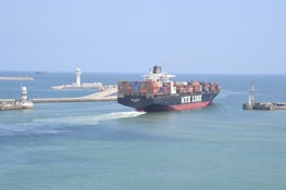 A large container ship labeled NYK LINE is navigating through a channel with breakwaters on either side. The ship is carrying numerous colorful shipping containers stacked on its deck. In the background, there are two lighthouses and another vessel can be seen on the open sea. The sky is clear and the water is calm, suggesting a peaceful maritime setting.