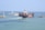 A large container ship labeled NYK LINE is navigating through a channel with breakwaters on either side. The ship is carrying numerous colorful shipping containers stacked on its deck. In the background, there are two lighthouses and another vessel can be seen on the open sea. The sky is clear and the water is calm, suggesting a peaceful maritime setting.