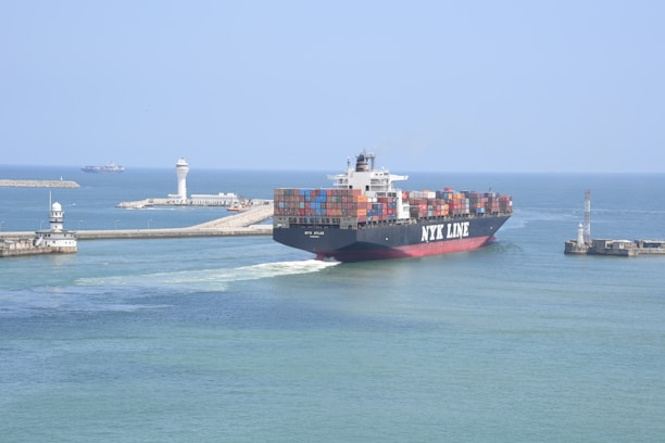 A large container ship labeled NYK LINE is navigating through a channel with breakwaters on either side. The ship is carrying numerous colorful shipping containers stacked on its deck. In the background, there are two lighthouses and another vessel can be seen on the open sea. The sky is clear and the water is calm, suggesting a peaceful maritime setting.