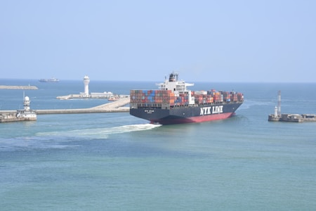 A large container ship labeled NYK LINE is navigating through a channel with breakwaters on either side. The ship is carrying numerous colorful shipping containers stacked on its deck. In the background, there are two lighthouses and another vessel can be seen on the open sea. The sky is clear and the water is calm, suggesting a peaceful maritime setting.