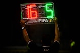 A person is holding up an electronic substitution board displaying the numbers 16 in red and 5 in green, typically used in football matches to indicate player changes.