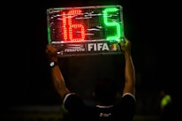 A person is holding up an electronic substitution board displaying the numbers 16 in red and 5 in green, typically used in football matches to indicate player changes.