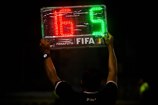 A person is holding up an electronic substitution board displaying the numbers 16 in red and 5 in green, typically used in football matches to indicate player changes.